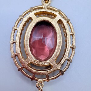 May include: A gold-toned pendant necklace with an oval, faceted, pink gemstone center. The gemstone is surrounded by a gold filigree design and small, pearl-like beads. The pendant has the word "AVON" stamped on it.