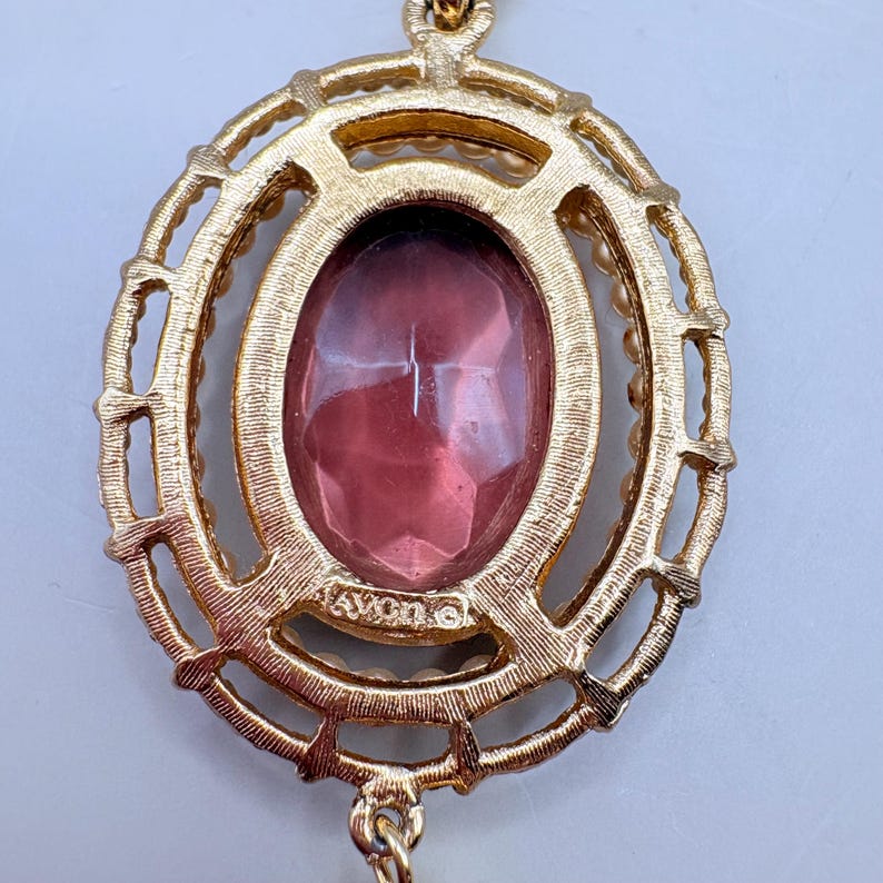 May include: A gold-toned pendant necklace with an oval, faceted, pink gemstone center. The gemstone is surrounded by a gold filigree design and small, pearl-like beads. The pendant has the word "AVON" stamped on it.