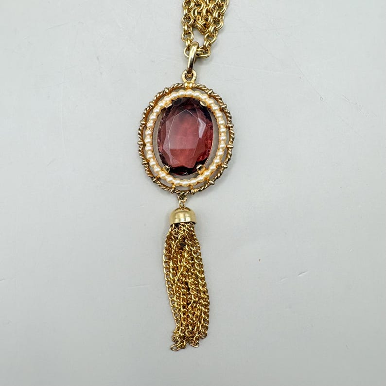 May include: Gold-tone necklace featuring a large, oval, burgundy-colored gemstone pendant. The pendant is surrounded by small, white pearls and a twisted gold rope design. A gold tassel hangs below the pendant, adding a touch of elegance.