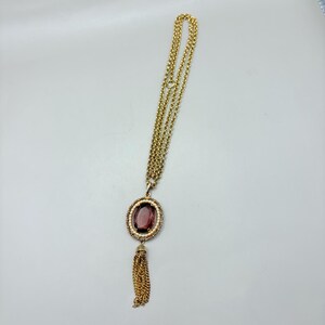 May include: Gold-toned necklace with a large oval pendant featuring a deep purple stone surrounded by small white beads. A gold tassel hangs below the pendant. The necklace chain is made of multiple strands.