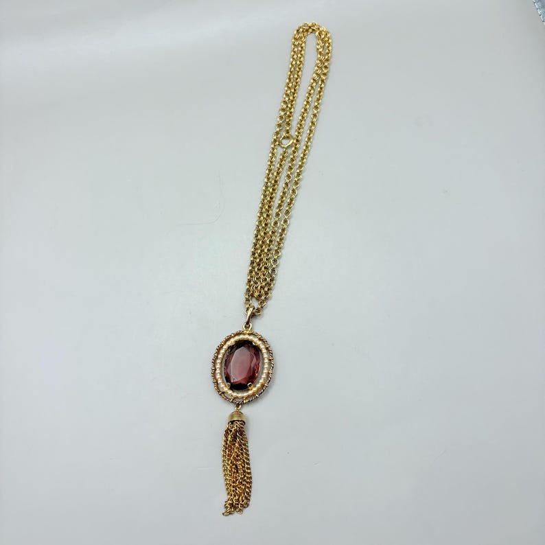 May include: Gold-toned necklace with a large oval pendant featuring a deep purple stone surrounded by small white beads. A gold tassel hangs below the pendant. The necklace chain is made of multiple strands.