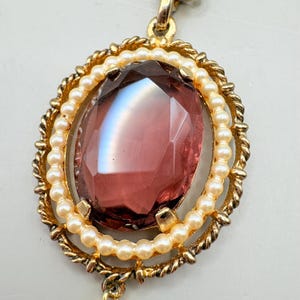 May include: A vintage pendant necklace featuring a large, faceted, oval-shaped, deep red gemstone. The gemstone is surrounded by a row of small, cream-colored faux pearls and a gold-toned, rope-textured frame. The pendant hangs from a delicate chain.