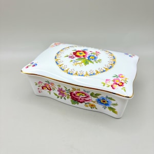 May include: A white porcelain jewelry box with a rectangular shape and a curved lid. The box is decorated with colorful floral patterns, including red, pink, yellow, and blue flowers. Gold trim accents the edges.