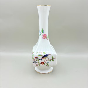 以下が含まれることがあります： 金色の縁と正方形のベースが付いた白いセラミック花瓶。花瓶には、ピンクの花と緑の葉が付いた枝に止まっている青い鳥の塗装デザインが施されています。花瓶は溝のあるデザインです。