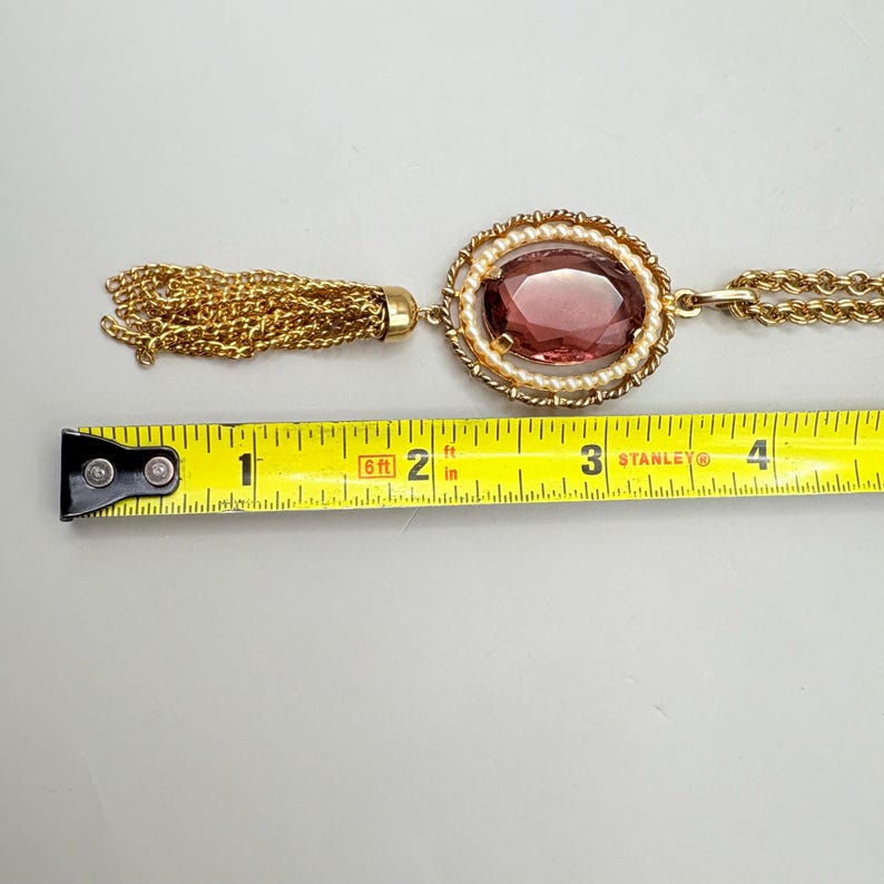 May include: Gold-tone necklace with a large oval, reddish-purple stone pendant, surrounded by faux pearls. The pendant is connected to a gold chain and a gold tassel. The necklace is approximately 3 inches long.