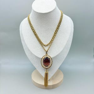 May include: A gold-toned necklace with a large oval, dark red pendant. The pendant is surrounded by small pearls and a gold frame. A gold tassel hangs below the pendant. The necklace is displayed on a white bust.