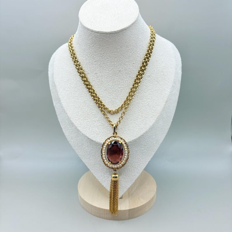 May include: A gold-toned necklace with a large oval, dark red pendant. The pendant is surrounded by small pearls and a gold frame. A gold tassel hangs below the pendant. The necklace is displayed on a white bust.