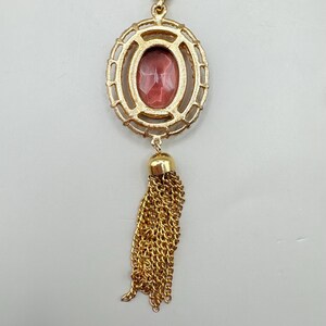 May include: Gold-toned pendant necklace featuring an oval-shaped, reddish-pink gemstone set within a decorative frame. A gold tassel hangs below the gemstone. The necklace is suspended from a delicate gold chain.