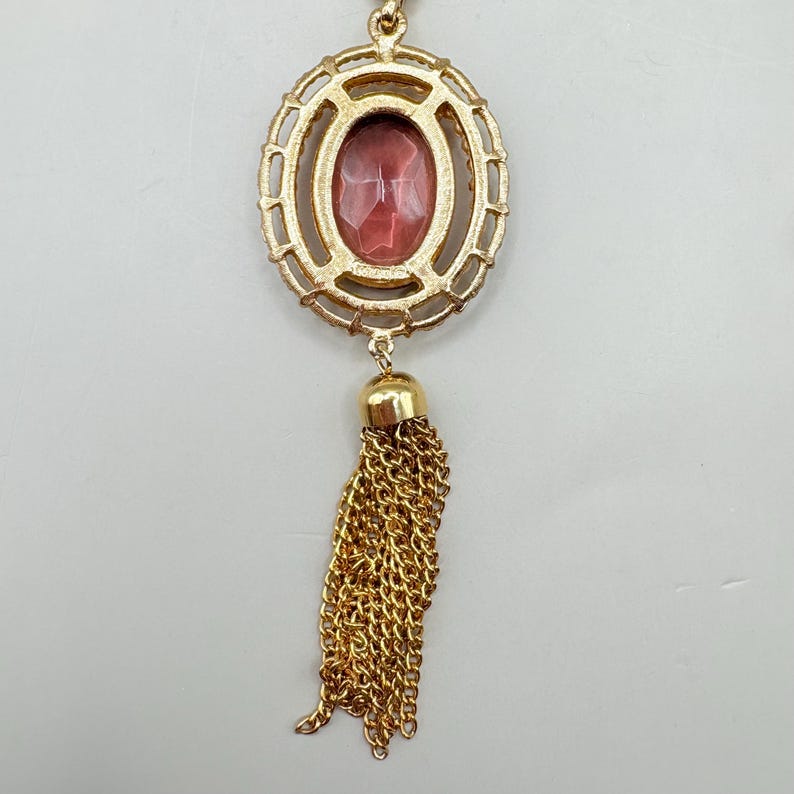 May include: Gold-toned pendant necklace featuring an oval-shaped, reddish-pink gemstone set within a decorative frame. A gold tassel hangs below the gemstone. The necklace is suspended from a delicate gold chain.