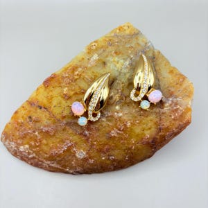 May include: A pair of gold-toned leaf-shaped earrings. Each earring features a leaf design with a row of small, clear crystals and two opal-colored stones. The earrings are displayed on a textured, tan-colored surface.