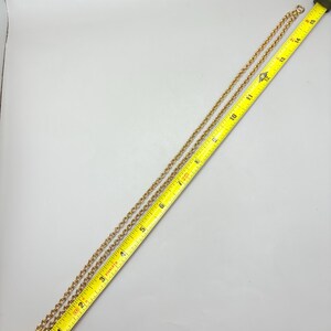 May include: A gold-colored chain necklace is laid out next to a yellow measuring tape. The chain is approximately 28 inches long. The necklace has a delicate, linked design. The measuring tape is angled across the image, providing a size reference.