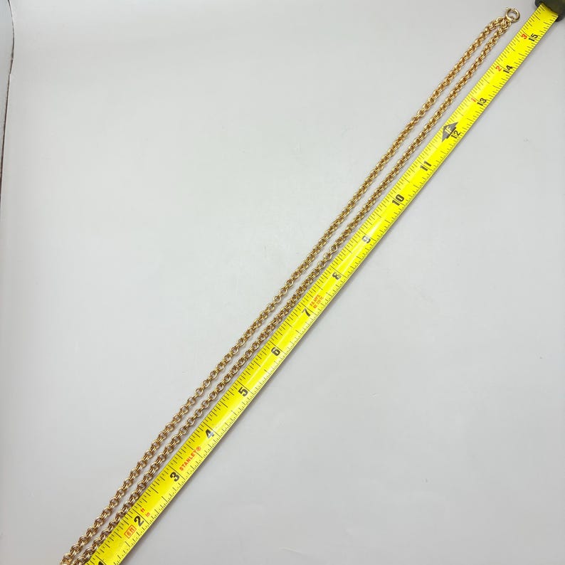 May include: A gold-colored chain necklace is laid out next to a yellow measuring tape. The chain is approximately 28 inches long. The necklace has a delicate, linked design. The measuring tape is angled across the image, providing a size reference.