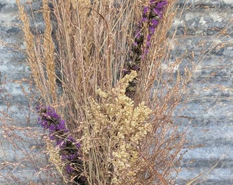 May include: A bouquet of dried grasses and flowers in shades of tan, beige, and purple, arranged in a round, dark gray metal bucket with two gold-toned handles. The arrangement features tall, thin stalks of grass and clusters of small blossoms.