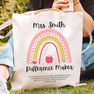 May include: Cream-colored tote bag with a rainbow and apple design. The text reads "Mrs. Smith Difference Maker" with a Bible verse below. The bag has two handles and is made of a sturdy fabric.
