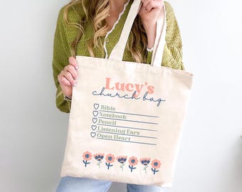 Può includere: Una borsa tote in tela beige con il testo "Lucy's church bag" in lettere rosa e blu. La borsa ha un elenco di articoli con cuori: Bibbia, Quaderno, Matita, Orecchie attente, Cuore aperto. La parte inferiore della borsa è decorata con una fila di fiori rosa.