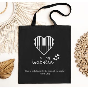 May include: Black tote bag featuring a white heart-shaped piano keyboard design. The name "isabella" is written in cursive below the heart, with musical notes to the right. The text "Make a joyful noise to the Lord, all the earth! Psalm 98:4" is printed at the bottom.