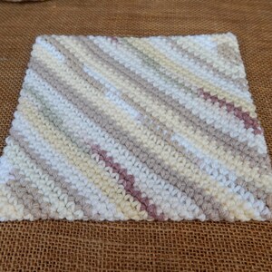 May include: A square, hand-knitted washcloth with a diagonal striped pattern. The stripes alternate between off-white, light gray, pale yellow, and a muted mauve color. The washcloth is made of soft yarn.