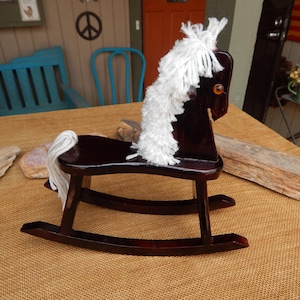 May include: A miniature wooden rocking horse with a dark brown finish. The horse features a white yarn mane and tail. The rocking horse is a classic toy, perfect for nursery decor or a collector's item.