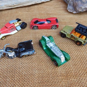 May include: A collection of toy cars, including a red hot rod, a red sports car, a green race car, a black motorcycle, and a green and tan jeep. The toys are on a brown surface.