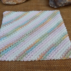May include: A hand-knitted square in pastel colors. The piece features diagonal stripes of pink, blue, green, and peach against a white background. The texture of the yarn is visible, suggesting a soft, handmade item. The item is likely a washcloth or a small blanket.