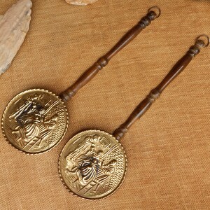 May include: Two antique brass bed warmers with wooden handles. The circular brass heads feature a detailed relief design. The handles are dark brown and have a ring at the end. The bed warmers are displayed on a textured, tan surface.