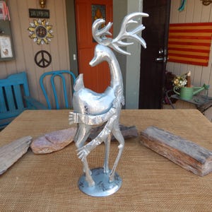 May include: A silver metal deer sculpture with large antlers and a circular base. The deer has a stylized design with outstretched arms and a textured surface. The sculpture is approximately 24 inches tall and is suitable for outdoor or indoor decor.