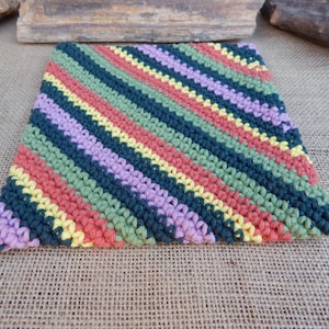 May include: A square, handmade crochet item with diagonal stripes in various colors. The stripes include shades of green, blue, pink, yellow, and purple. The item is likely a washcloth or trivet.