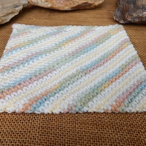 May include: A square, hand-knitted washcloth with a diagonal striped pattern. The cloth features pastel colors including white, yellow, blue, green, and peach. The washcloth is made of soft yarn and is suitable for gentle cleansing.