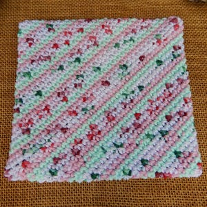 May include: A square, handmade crochet washcloth with a diagonal striped pattern. The stripes alternate between light pink, light green, and white, with small red and green floral accents throughout. The washcloth is displayed on a brown burlap surface.