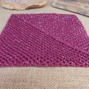 May include: A square, hand-crocheted coaster in a deep magenta color with silver metallic thread woven throughout. The coaster is made with a textured stitch and sits on a neutral burlap surface.