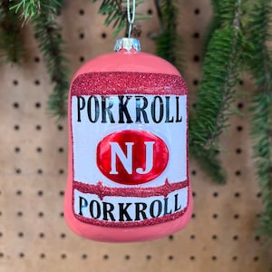 May include: A pink Christmas ornament shaped like a pork roll can. The ornament has red glitter accents and the words "PORKROLL" and "NJ" in a red oval. A silver string is attached for hanging.