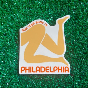 May include: White sticker with an orange outline and the text "SUN YOUR BUNS IN PHILADELPHIA". The sticker features a stylized illustration of a person's legs in a sitting position.