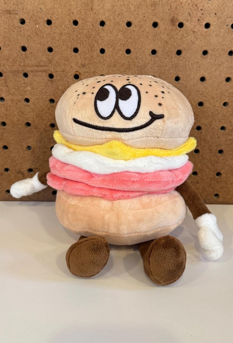 Puede incluir: Un peluche con forma de s&aacute;ndwich sonriente. Tiene un bollo marr&oacute;n claro, carne rosa, queso blanco y huevo amarillo. El juguete tiene brazos y pies marrones, con guantes blancos. Tiene ojos grandes y de dibujos animados.