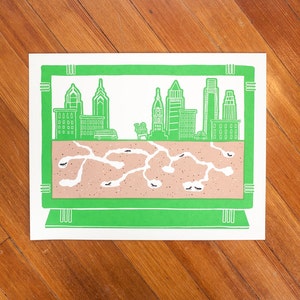 May include: A green framed illustration of a city skyline with a brown ant colony underneath. The city skyline includes several buildings and a car. The ant colony is made up of tunnels and chambers.