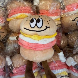 Puede incluir: Juguete de peluche con forma de hamburguesa con una cara sonriente, brazos y piernas. La hamburguesa tiene un pan tostado, una hamburguesa rosa, queso amarillo y salsa blanca. El juguete est&aacute; rodeado de otras hamburguesas de peluche similares.