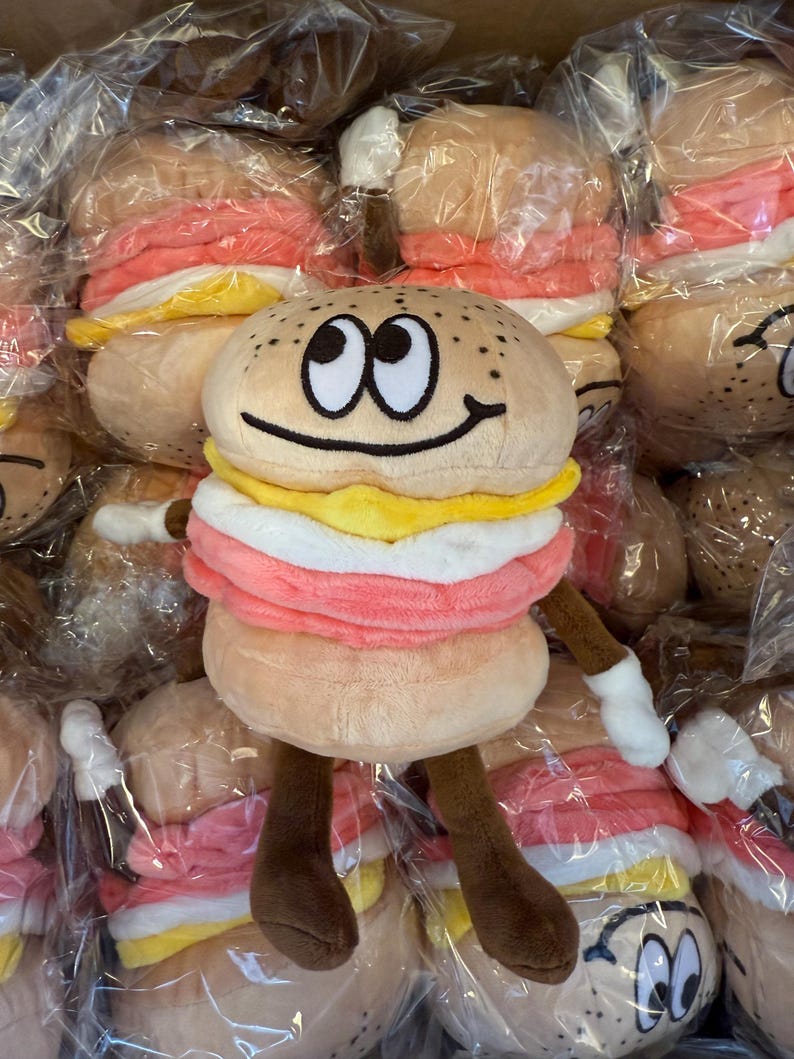 Puede incluir: Juguete de peluche con forma de hamburguesa con una cara sonriente, brazos y piernas. La hamburguesa tiene un pan tostado, una hamburguesa rosa, queso amarillo y salsa blanca. El juguete est&aacute; rodeado de otras hamburguesas de peluche similares.