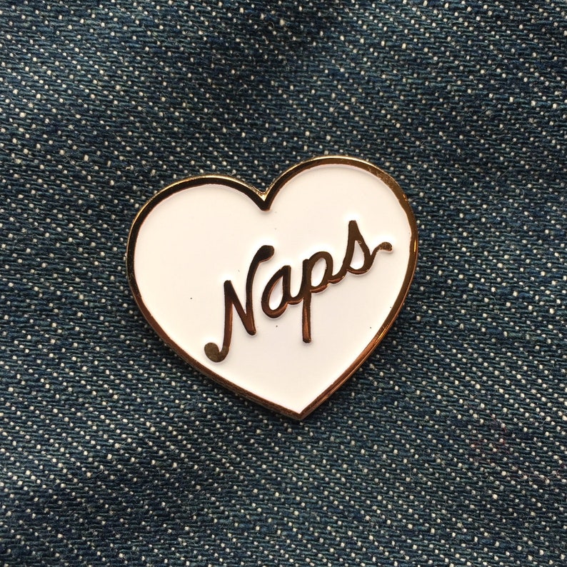 Pode incluir: Pin de esmalte branco em forma de cora&ccedil;&atilde;o com contorno dourado e a palavra "Naps" escrita em letras cursivas douradas.