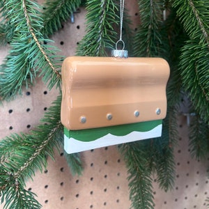 May include: A tan and green ceramic Christmas ornament shaped like a squeegee. The ornament has a white trim and three silver metal accents.