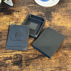 Puede incluir: Juego de tres estuches rectangulares de color gris oscuro. El juego incluye una base, una tapa y un inserto más pequeño. La tapa tiene una superficie lisa, mientras que la base tiene un interior texturizado. La parte frontal de la tapa tiene el texto "SCdesignENG" y un gráfico.