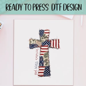 May include: A ready to press DTF design featuring a cross shape composed of patchwork fabric patterns including American flag stripes, stars, and camouflage. The text 'god bless america' is written in a red script font along the left side.