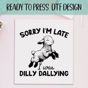May include: A white square with the text "READY TO PRESS DTF DESIGN" at the top. The design features a jumping lamb with the text "SORRY I'M LATE I WAS DILLY DALLYING" in black. The design is suitable for apparel.