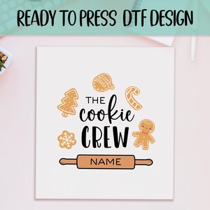 May include: White square with a "READY TO PRESS DTF DESIGN" banner at the top. The design features the text "THE cookie CREW" with gingerbread cookie illustrations and a rolling pin. The word "NAME" is below the rolling pin.
