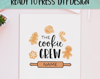 May include: White square with a "READY TO PRESS DTF DESIGN" banner at the top. The design features the text "THE cookie CREW" with gingerbread cookie illustrations and a rolling pin. The word "NAME" is below the rolling pin.