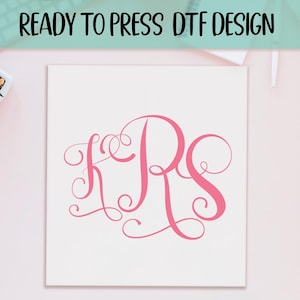May include: A white square with the text "READY TO PRESS DTF DESIGN" at the top. The design features the pink cursive letters K, R, and S in a monogram style. The background is a light pink surface.