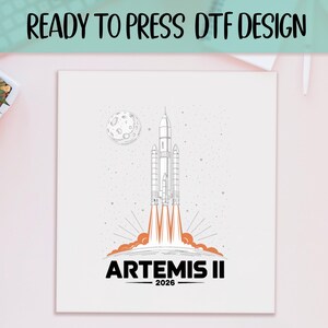 May include: White square print featuring a rocket ship launching into space, with a moon in the background. The design includes the text "READY TO PRESS DTF DESIGN" at the top and "ARTEMIS II 2026" at the bottom in black and orange.