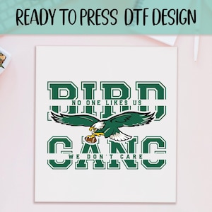 May include: White square with a green and white design. The design features an eagle holding a football, with the words "DIDD", "NO ONE LIKES US", and "GANG WE DON'T CARE" in a collegiate font. The words "READY TO PRESS DTF DESIGN" are at the top.