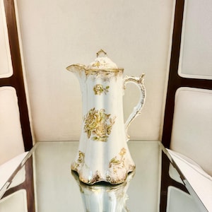 May include: A tall, off-white porcelain pitcher with a lid, featuring a delicate floral pattern of yellow and brown roses. The pitcher has a scalloped base, a curved handle, and gold-toned trim accents along the rim, handle, and base edges.