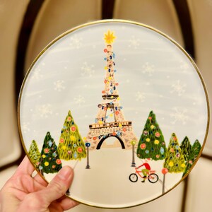 May include: A round decorative plate with a gold rim. The plate features a whimsical illustration of the Eiffel Tower decorated with Christmas lights, surrounded by Christmas trees and snowflakes. A person rides a bicycle in the foreground.