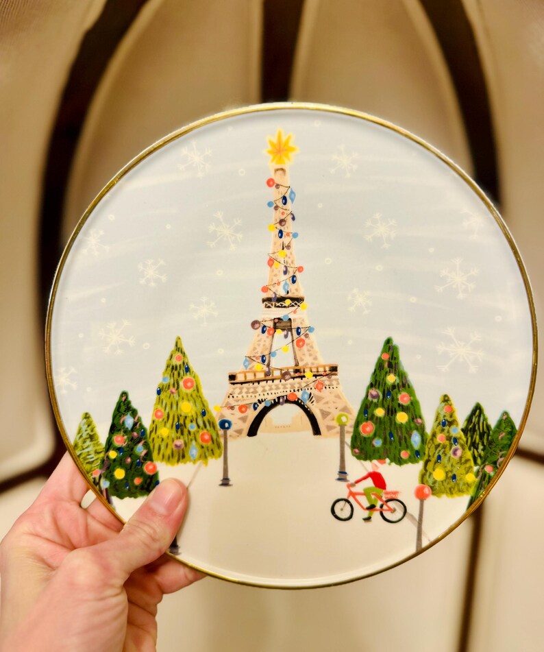 May include: A round decorative plate with a gold rim. The plate features a whimsical illustration of the Eiffel Tower decorated with Christmas lights, surrounded by Christmas trees and snowflakes. A person rides a bicycle in the foreground.