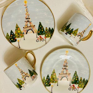 May include: A set of two plates and two mugs with a winter scene featuring the Eiffel Tower decorated with lights, Christmas trees, and a cyclist. The plates and mugs have a gold rim and handle.